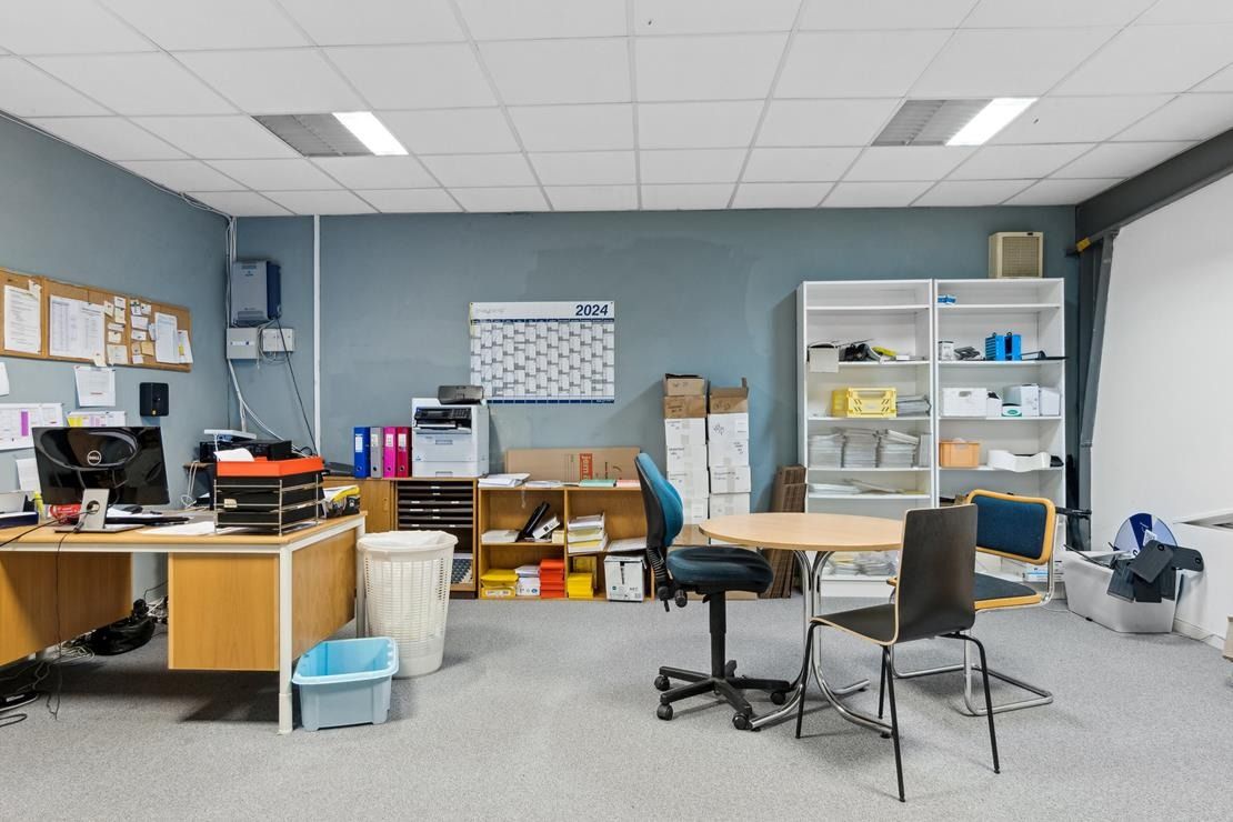 Kontor med skrivebord, kalender og flere hylder. Bord med computer og papirkurv samt et runde bord med stol.