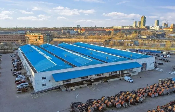 En stor lagerbygning med blå tag og parkeringsplads med mange biler og lastbiler foran bygningen.