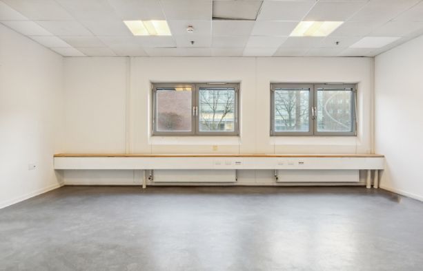 Rummelige lokaler med højt gulv og hvide vægge og to store vinduer med udsigt til bygning og træer