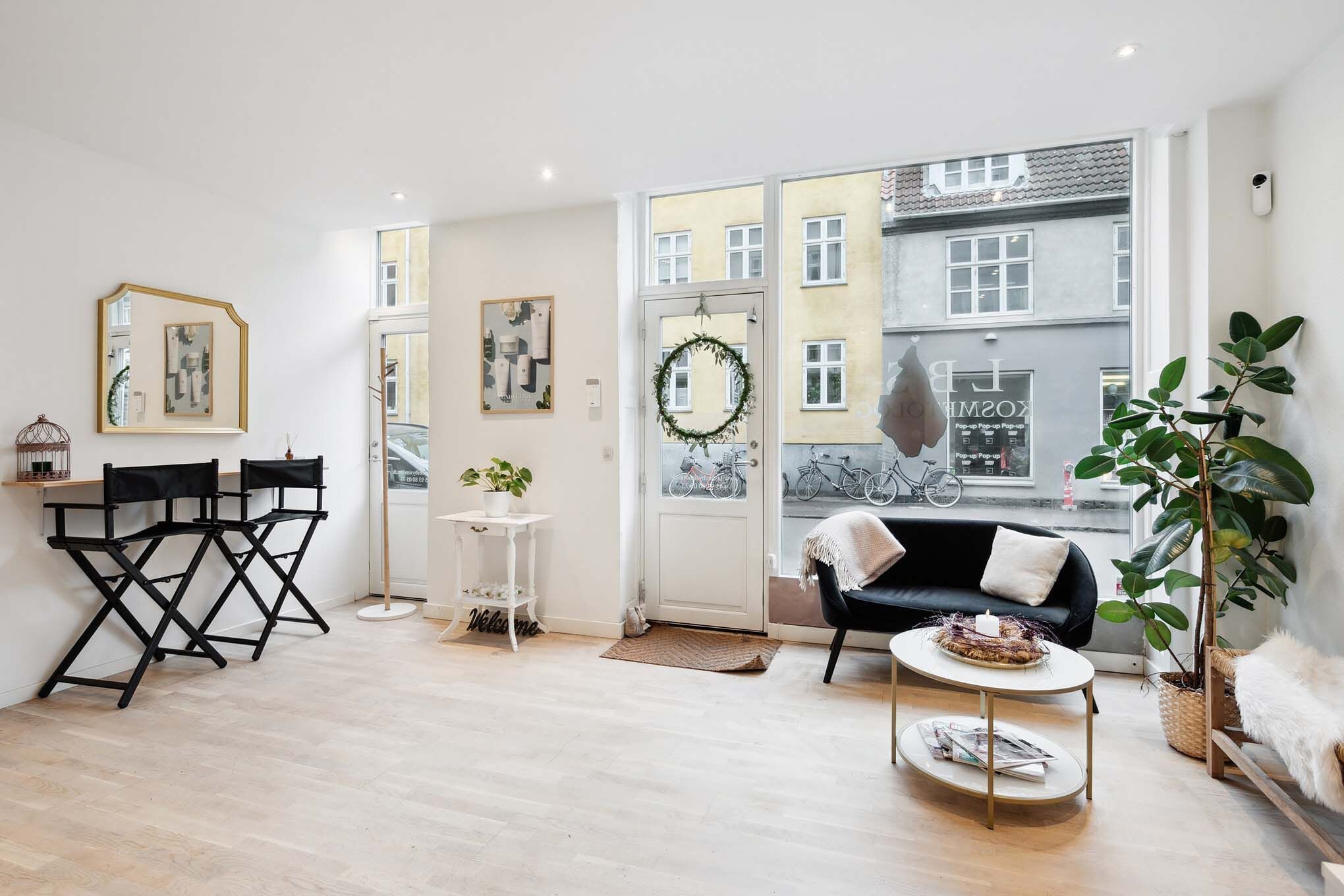 Minimalistisk stue med hvide vægge, trægulv, to højsæde, sofa med pude og bord med plante samt vindue med udsigt til gaden