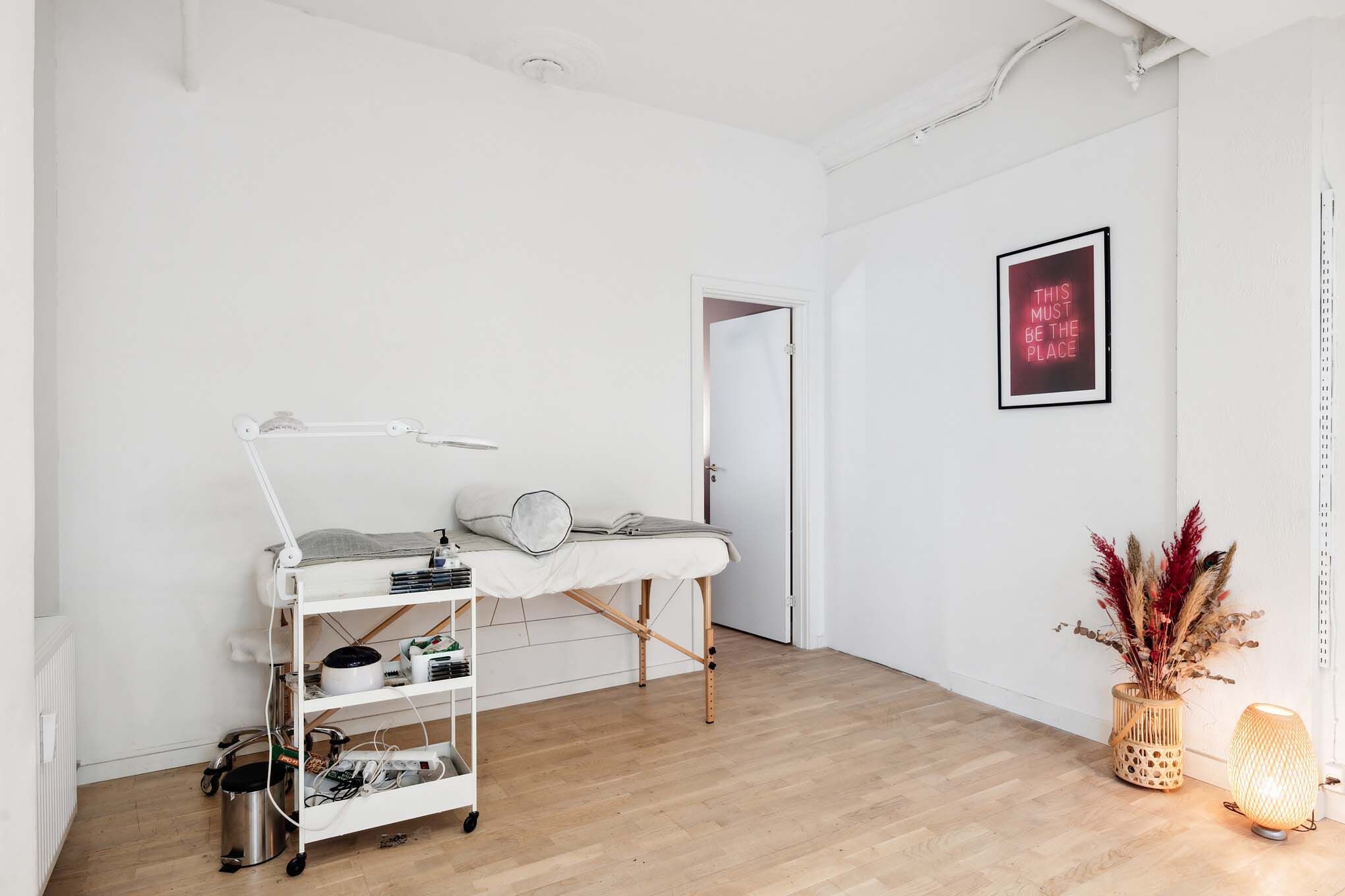 Kosmetologpraksis med massagebord, håndklæder, massagelys, plante og lampe i hjørnet af en hvid stue med en neonskiltet på