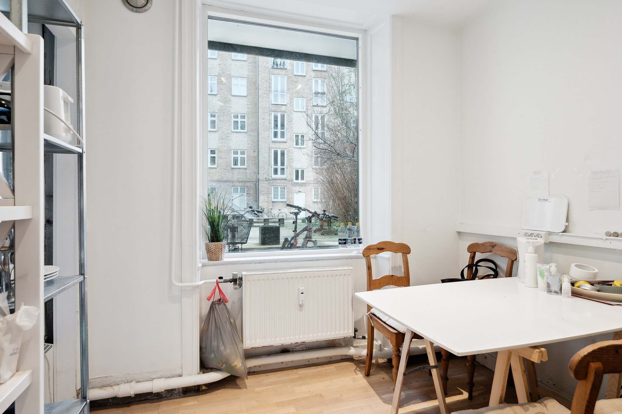 En moderne hvid stue med trægulv, hvidt bord, træstole og plante i vindueskarme, radiator og kludposer