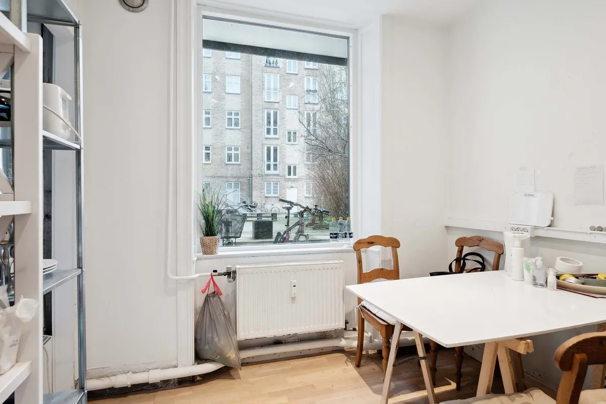 En moderne hvid stue med trægulv, hvidt bord, træstole og plante i vindueskarme, radiator og kludposer