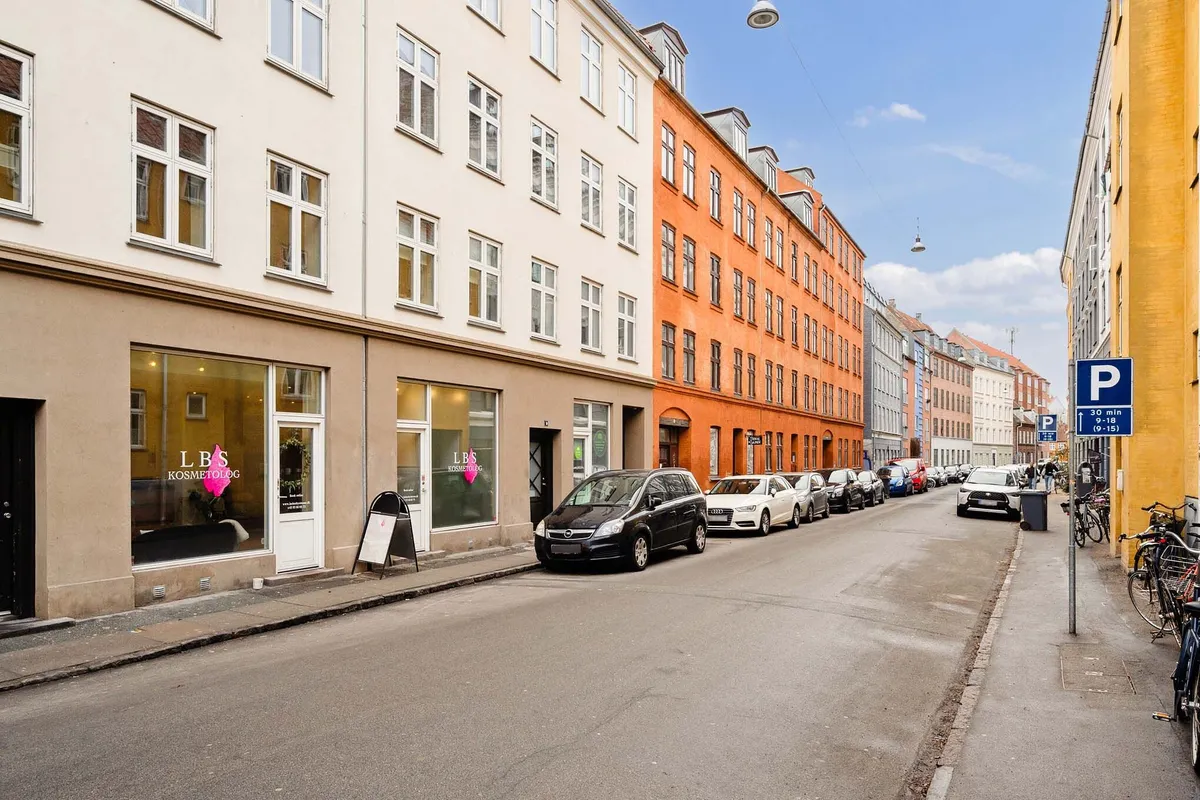 Store bygninger med butikker og parkerede biler, vejspærring og cykler stående langs fortov, lys og skyer, gul og orange