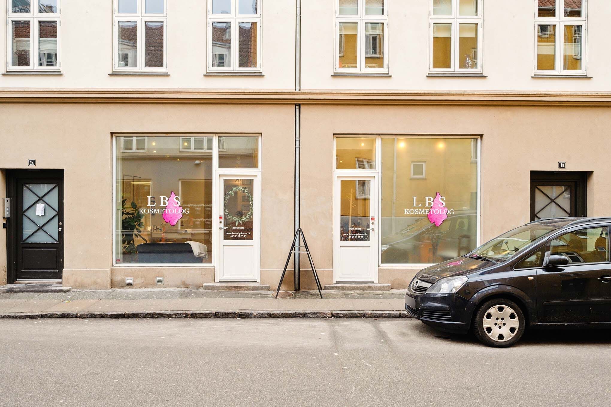 Kosmetologklinikken LBS ligger på gadekant og har to hvide døre med pink pige og tekst