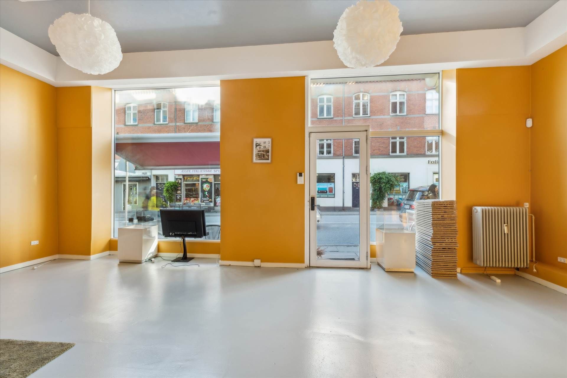 Et lyst og tomt lokale med gule vægge og hvide loftslamper. Rummet har gulv og vinduer mod gaden og en dør mod køkken.