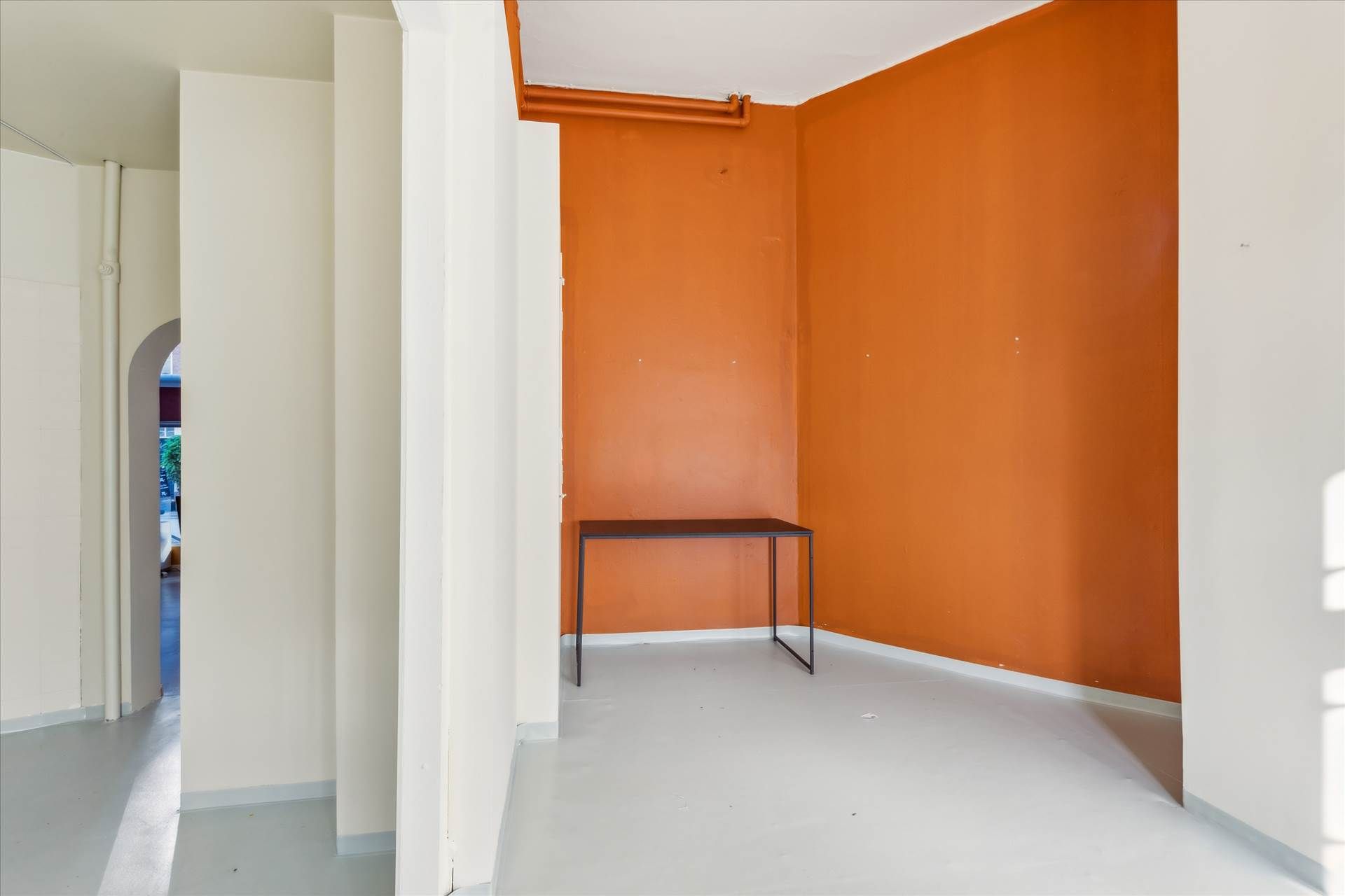 En tømt stue med orange væg, hvid loft og gulv samt et lille sort bord med ben i metal