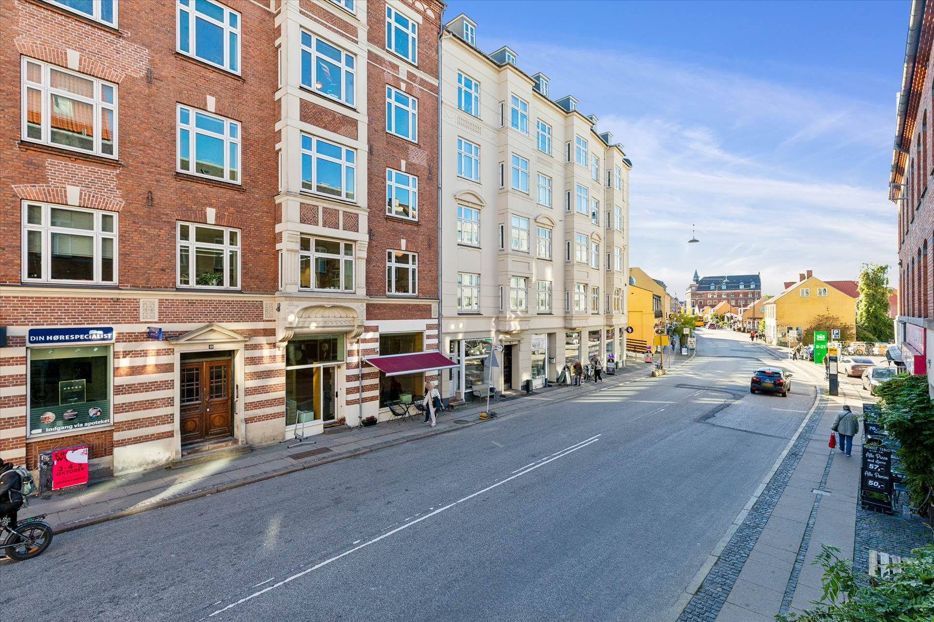 En tom gade med forretninger på hjørner og mennesker, der går på fortovet, og biler parkeret langs vejen