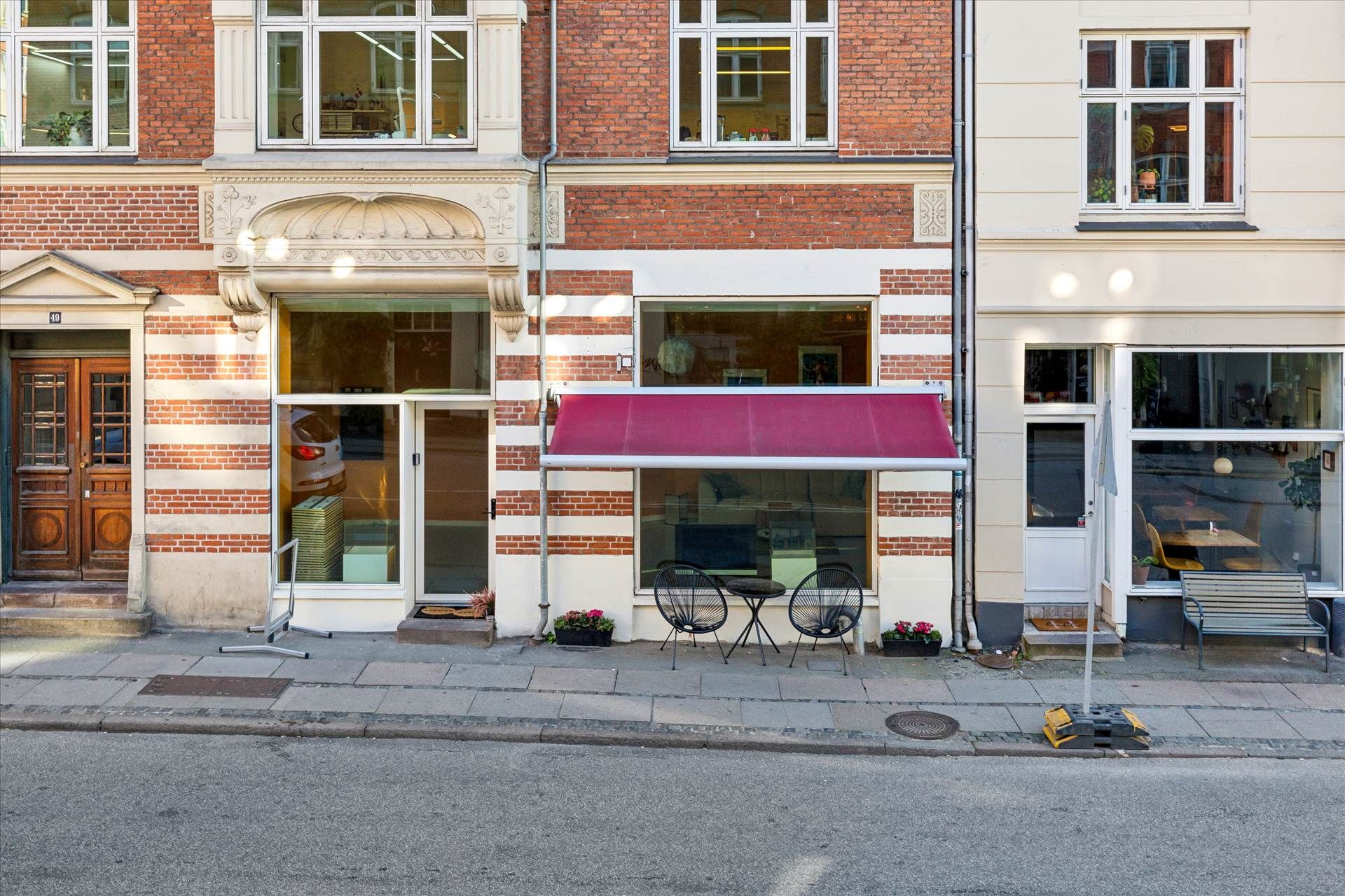 En forretning med rødt tag og to stole og et lille bord udenfor på gågaden, med flere butikker i baggrunden