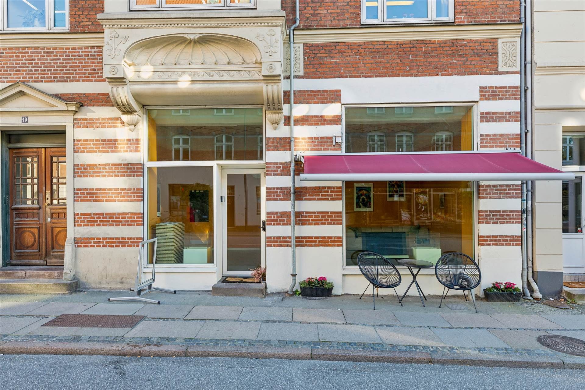 Butiksfront med rød markise, stole og bord, blomster i potte og lyseruder med udsigt til gaden