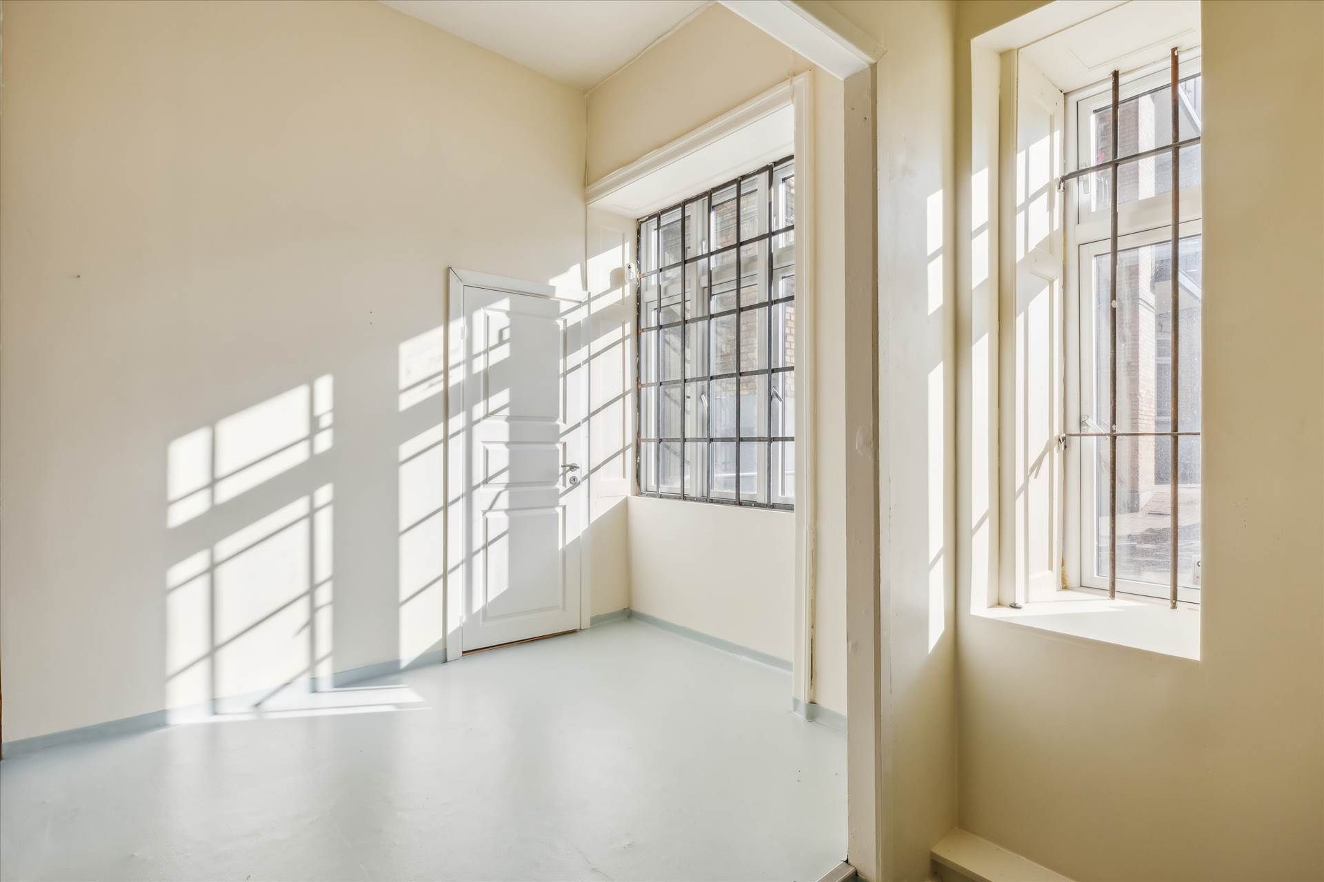 En tom kontorvæg med to vinduer og en hvid dør, hvor dagslys strømmer ind og kaster skygger på væggen