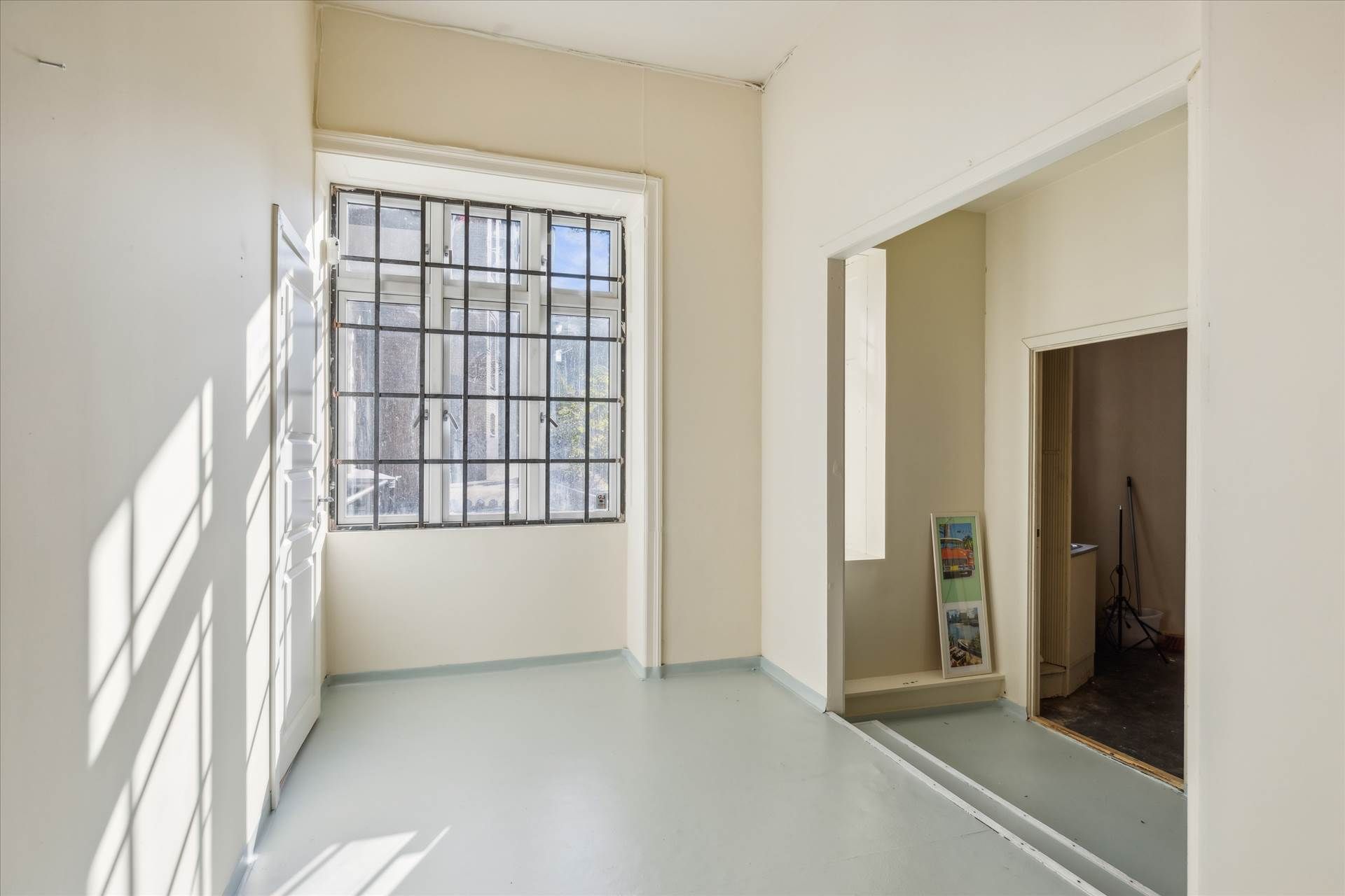 En stor, tom stue med hvide vægge og lyse skyggespil, der falder på væggen fra det store vindue. I vinduet ses en bygning