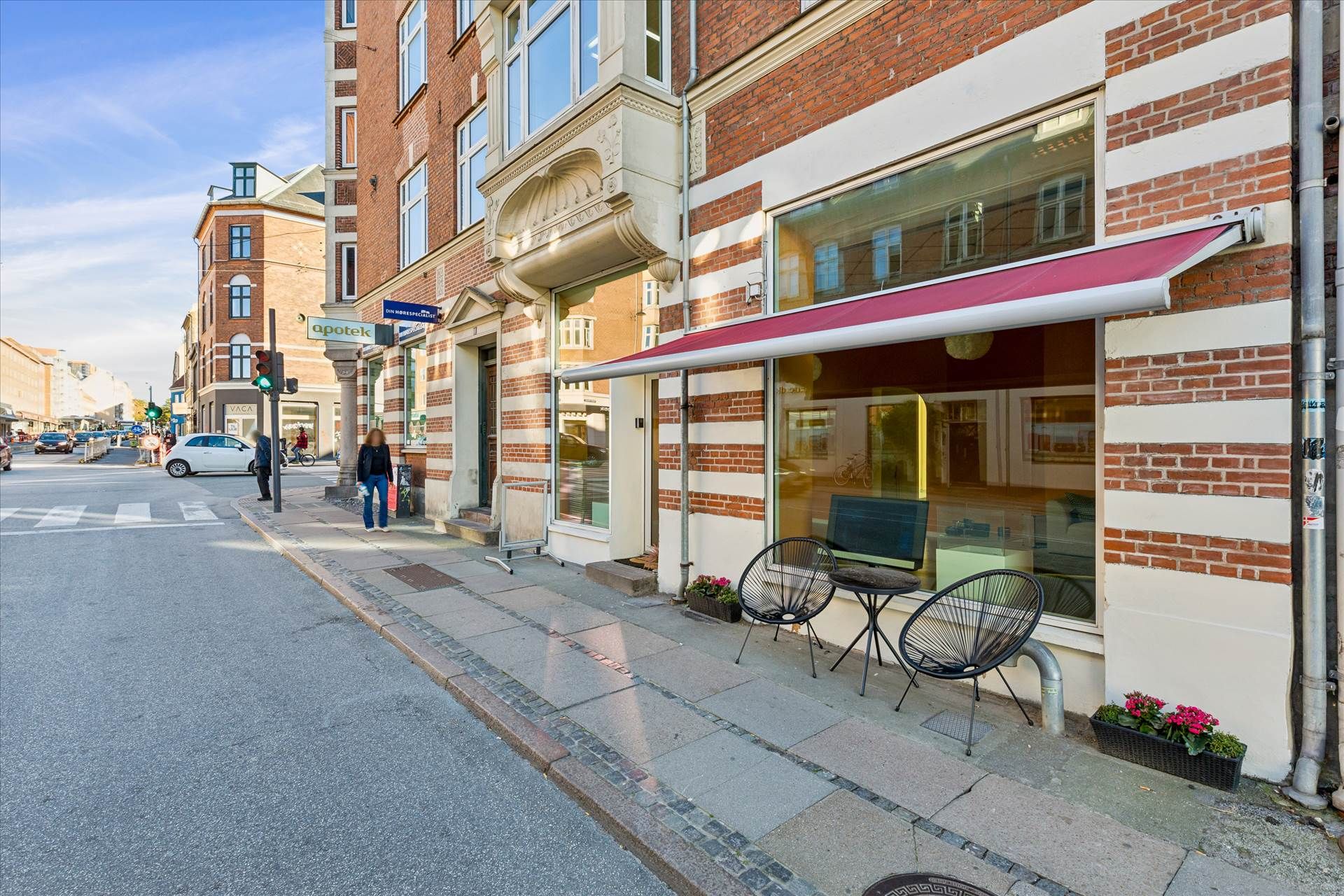 Butik med rød markise, hvor to mennesker går forbi, og der er en bil på vejen