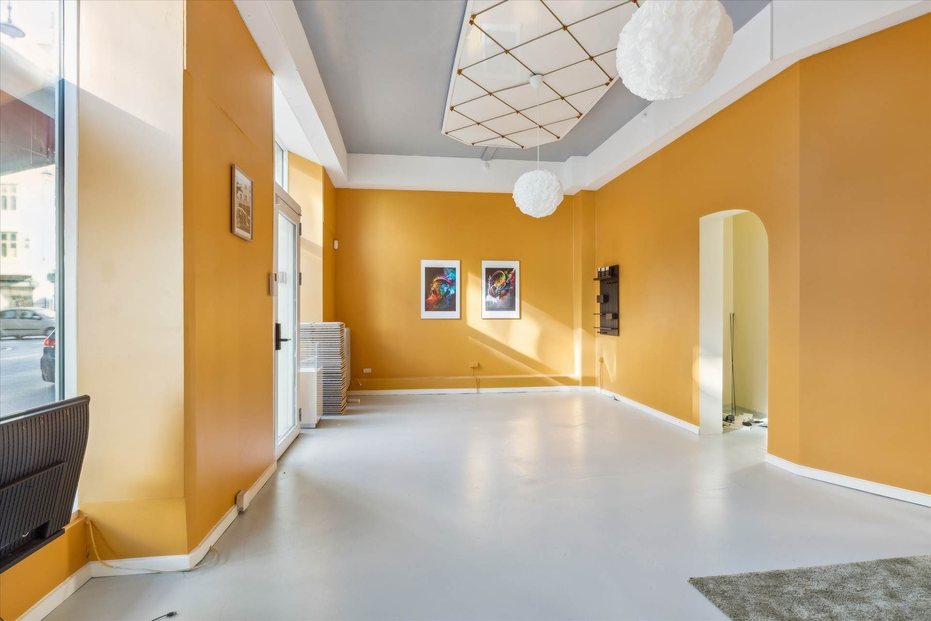 Kort og slank erhvervslejlighed med højt til loftet og lysegul væg og hvid gulv. Billedet viser to billeder hængende på