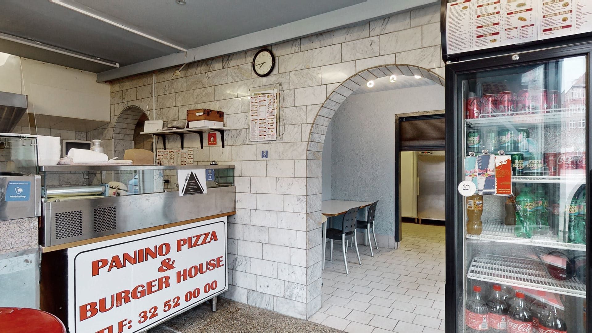 En pizza- og burgerrestaurant med et skab med sodavand og andre drikkevarer samt en udstilling med menukort på væggen