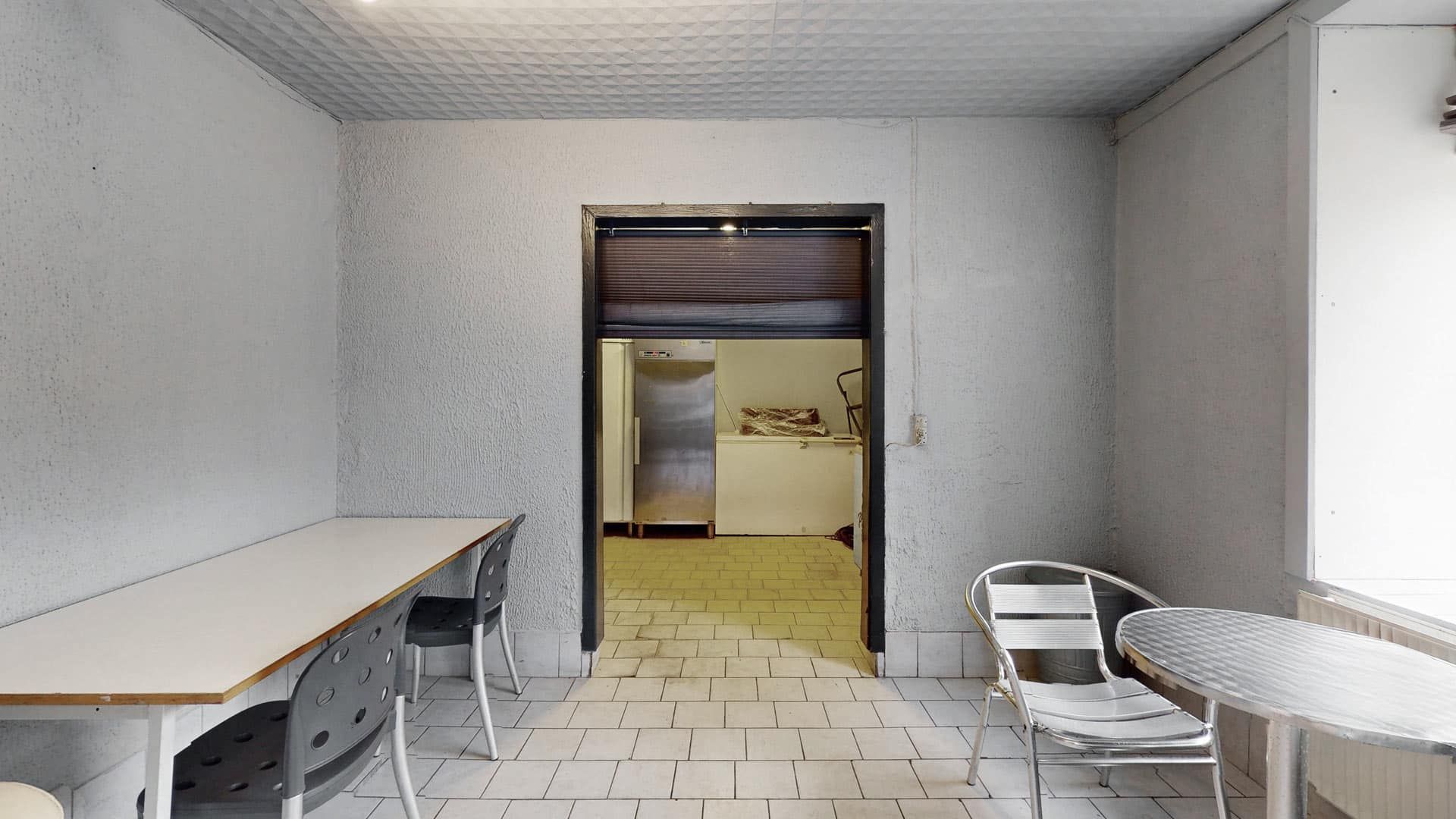 En tom og ren stue med to runder træborde og fire stole, hvor det er mørkt og der er et køleskab i baggrunden