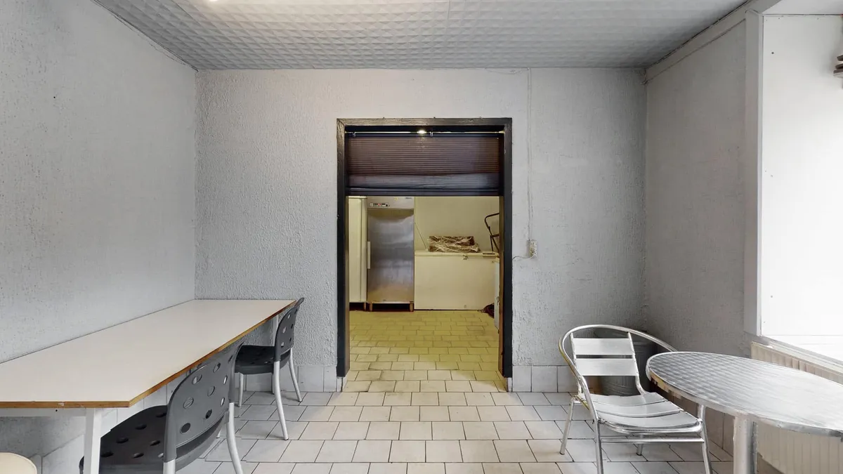 En tom og ren stue med to runder træborde og fire stole, hvor det er mørkt og der er et køleskab i baggrunden