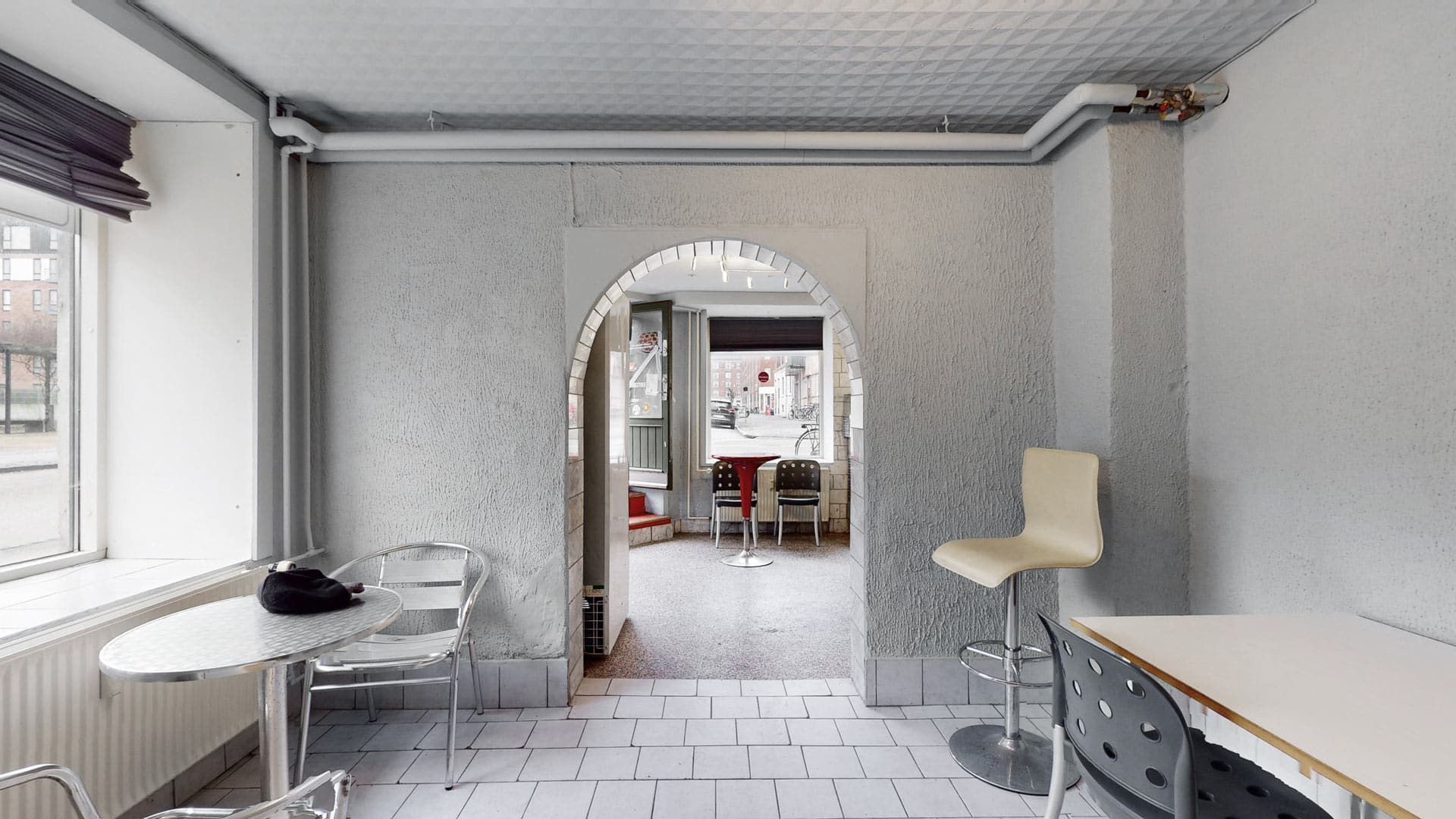 Værelse med lysegul væg og hvidt loft samt flere stole og borde med en åbning til en anden del af lokalet