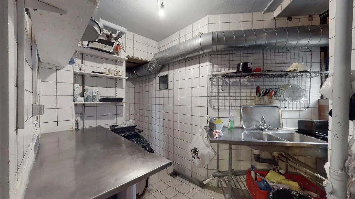 Et køkken med en stor metalarbejdsbord, to vasker med en håndvask og et hultag med ventiler, og en metalreol med gryder og