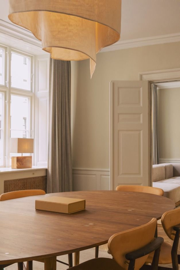 Rundt spisebord med lyse, runde stole i lysebrunt læder og træ. Moderne lysekron og gulvmøbler i lyse nuancer. Vindue med