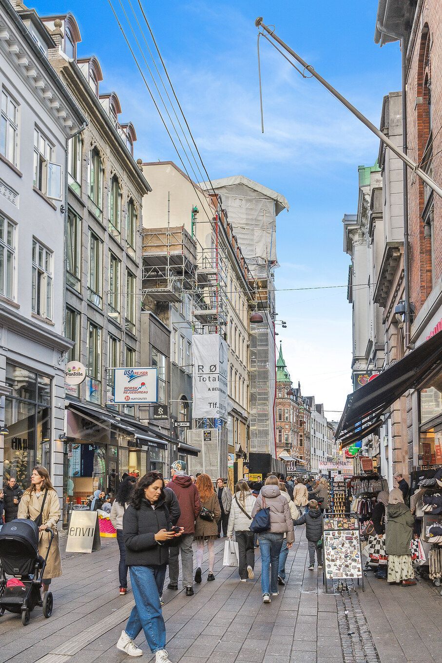 Mennesker går i en gågade med bygninger og butikker på begge sider, sammen med en kvinde der kører en barnevogn