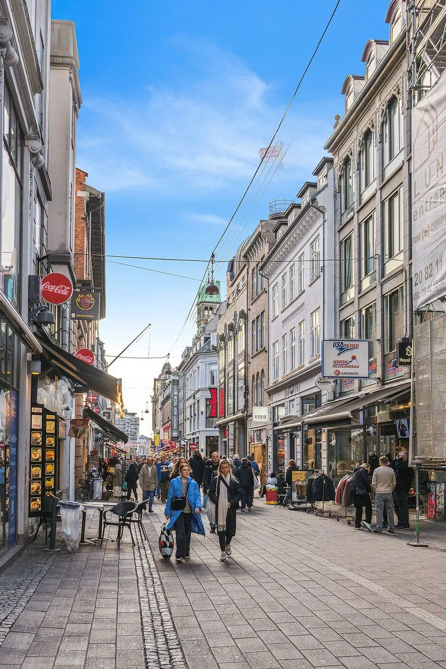Folk vandrer på et torv med små butikker og huse i baggrunden, udstyret med skilte og reklamer