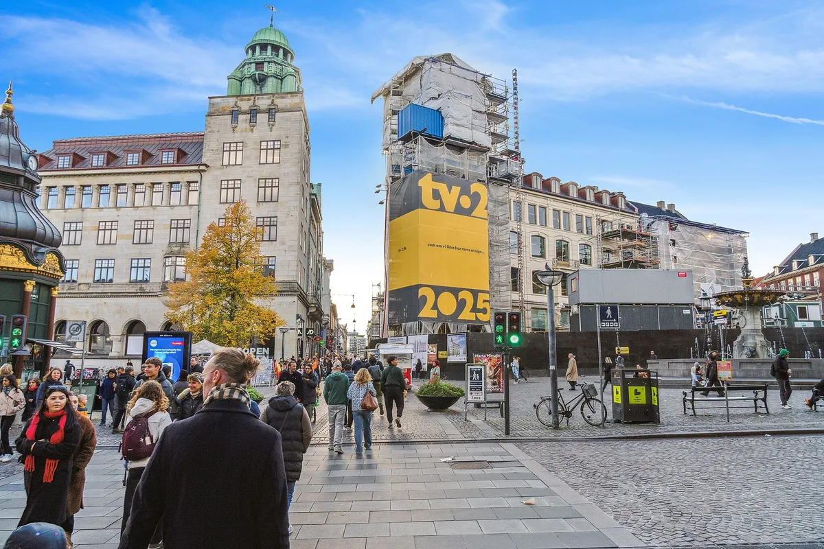 Mennesker strømmer gennem byens gade med tv2-skilt og 2025-annonce på facaden, mens folk går forbi og parkerede cykler og