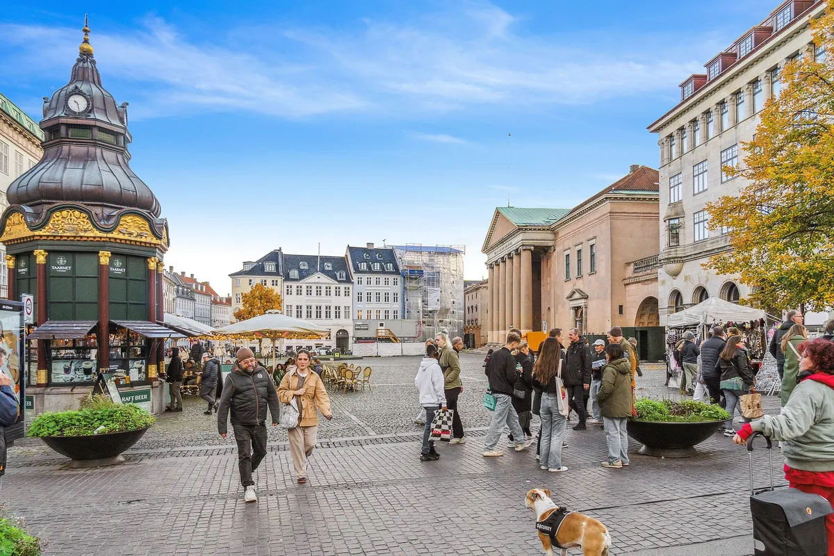 Mennesker vandrer gennem en åben gade med flere bygninger, pavilloner og gamle bygninger i baggrunden, og det er en solrig