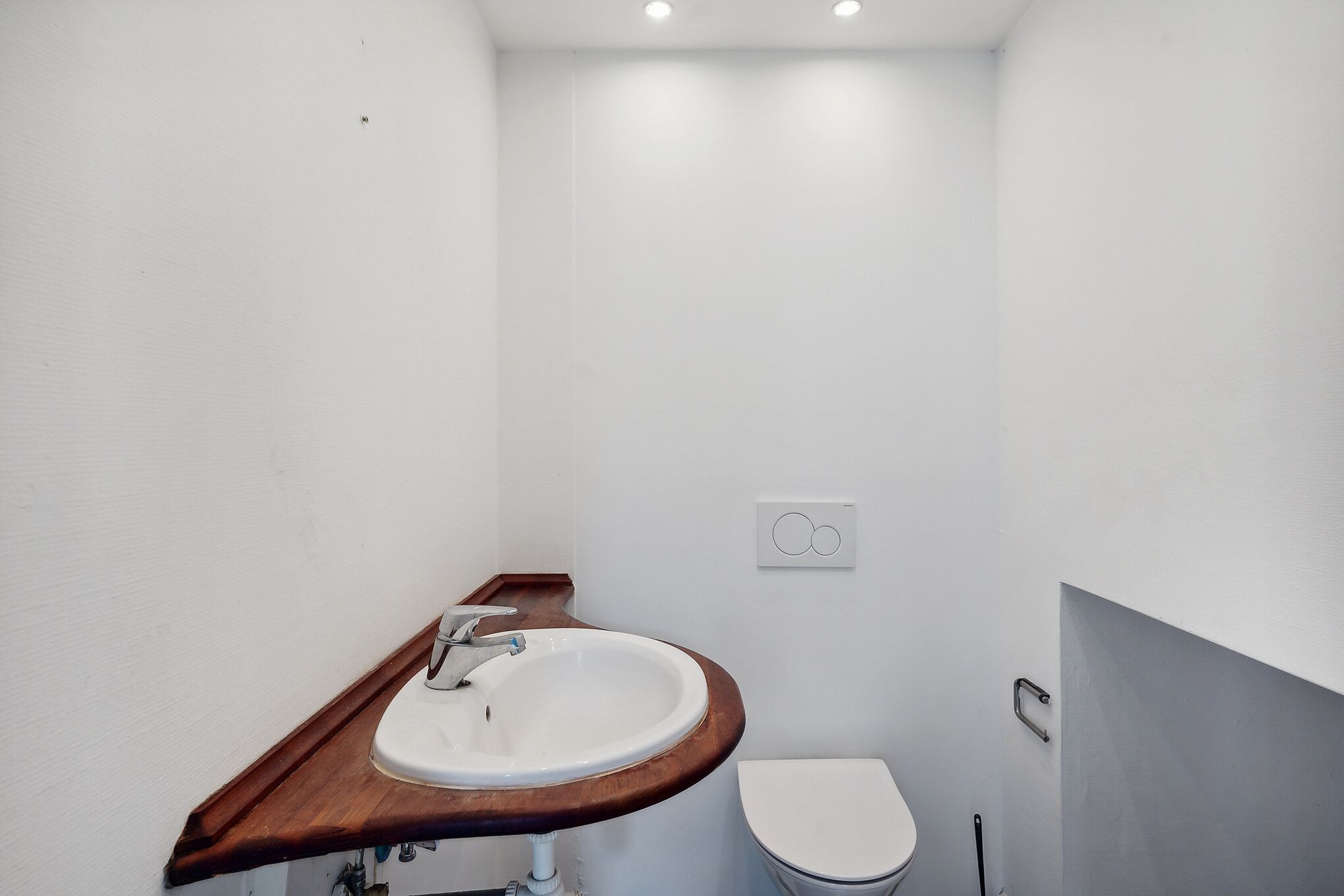 En lille og hvidmalet toiletrum med toilet, håndvask og et simpel udvendigt skab med håndtag og ventiler