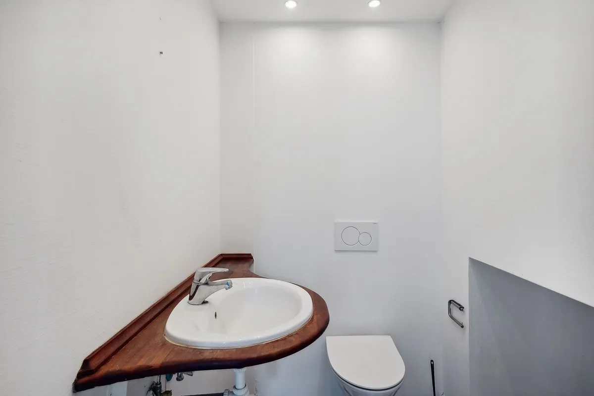 En lille og hvidmalet toiletrum med toilet, håndvask og et simpel udvendigt skab med håndtag og ventiler