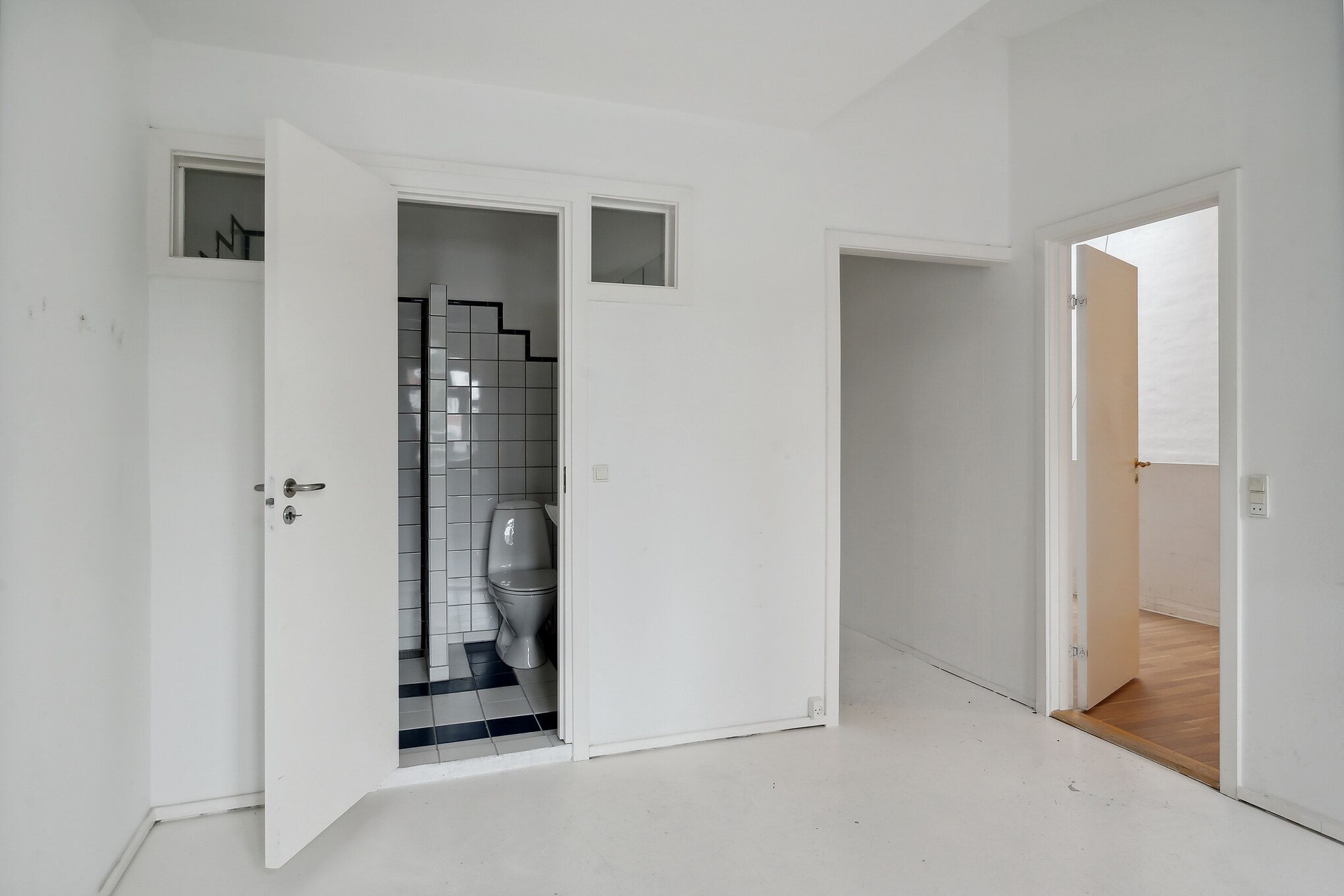 Værelse med hvid væg og højloft og toilet med hvid toiletblok og hvide kakelflader