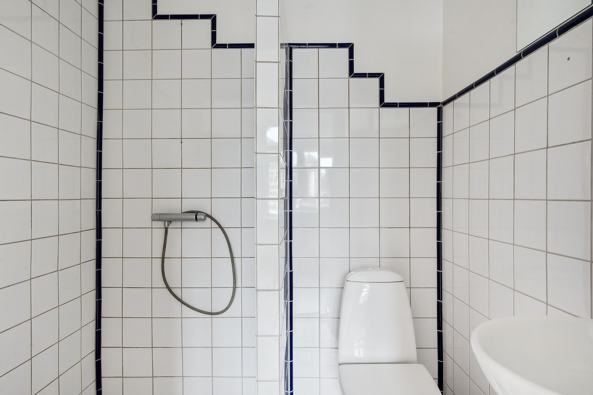 En hvid badeværelsesmøbel med toilet, vask og hængende hånddusch. Badrummet har hvide fliser med blå accenter.