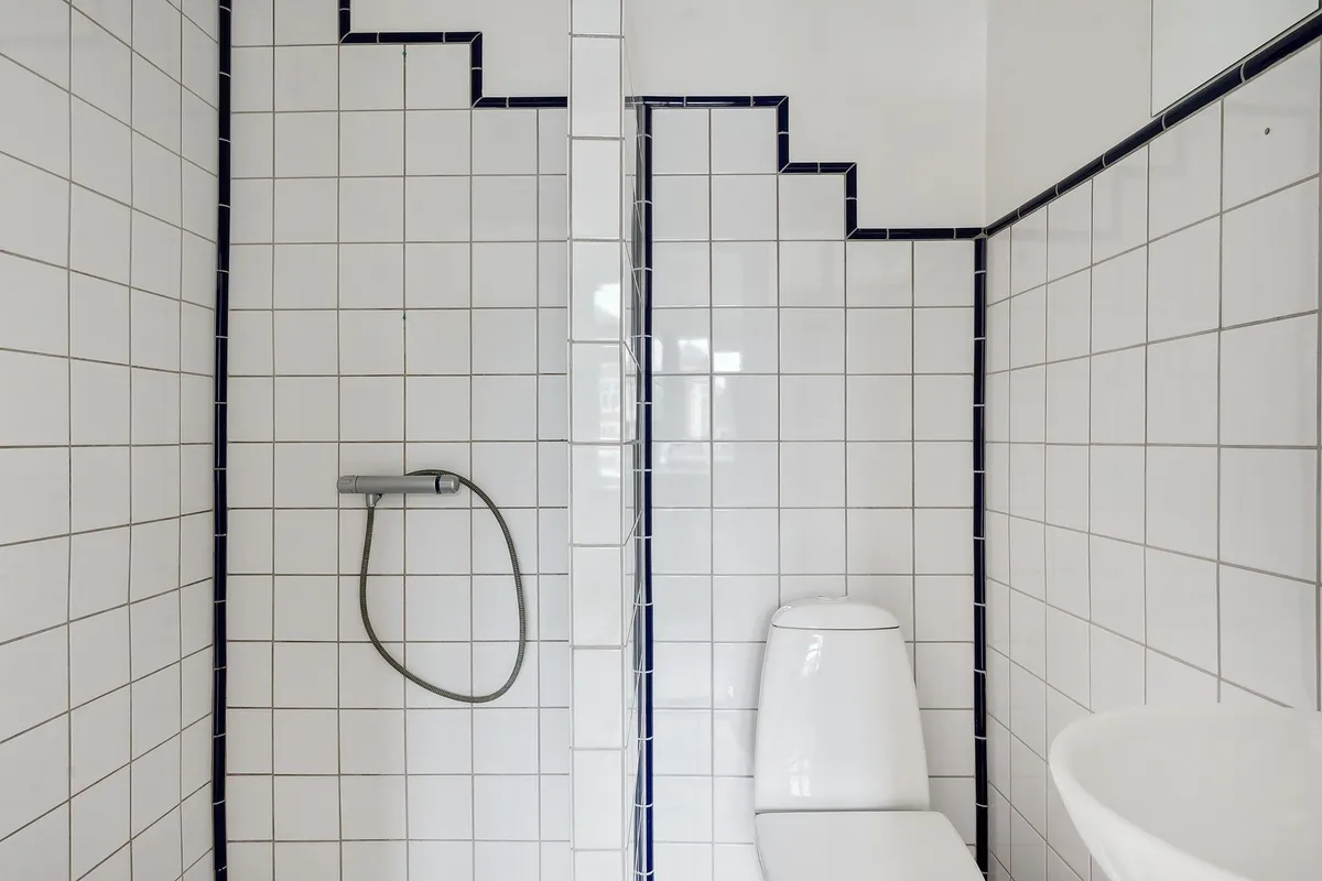 En hvid badeværelsesmøbel med toilet, vask og hængende hånddusch. Badrummet har hvide fliser med blå accenter.