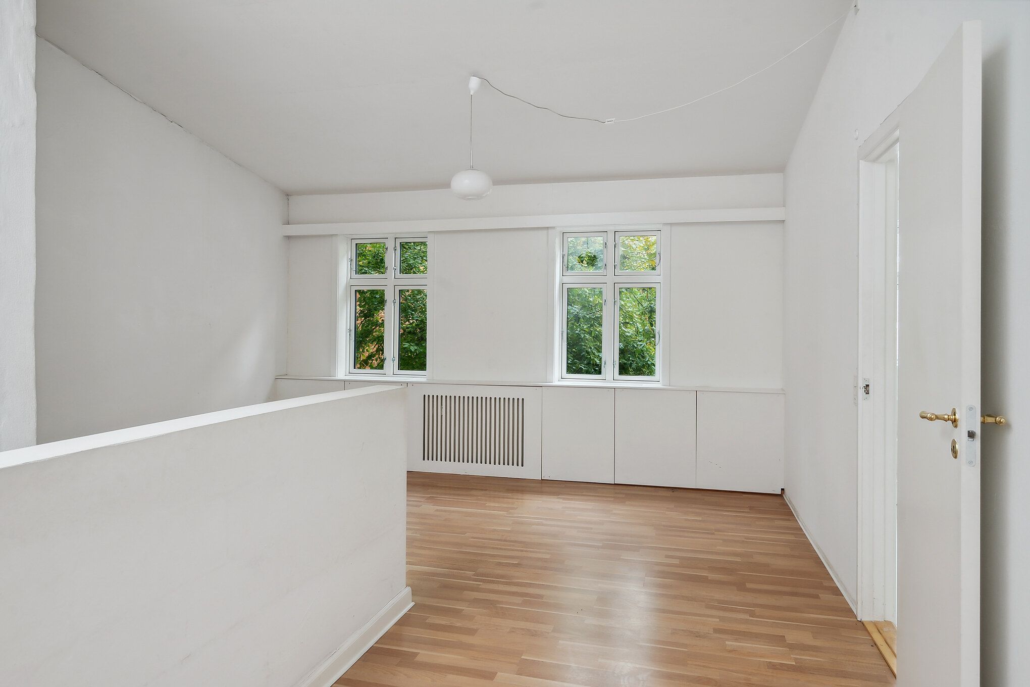 Et værelse med hvide vægge og gulv samt to vinduer med udsigt til grønne træer udenfor