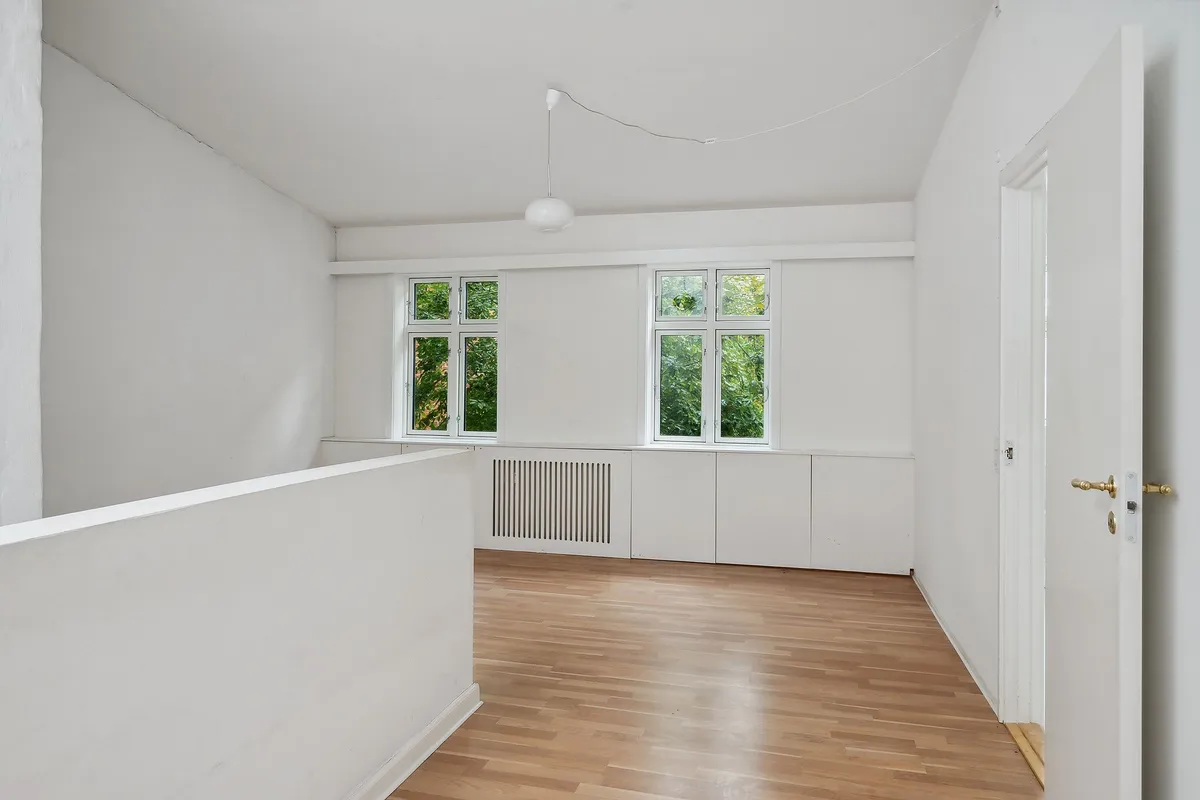 Et værelse med hvide vægge og gulv samt to vinduer med udsigt til grønne træer udenfor