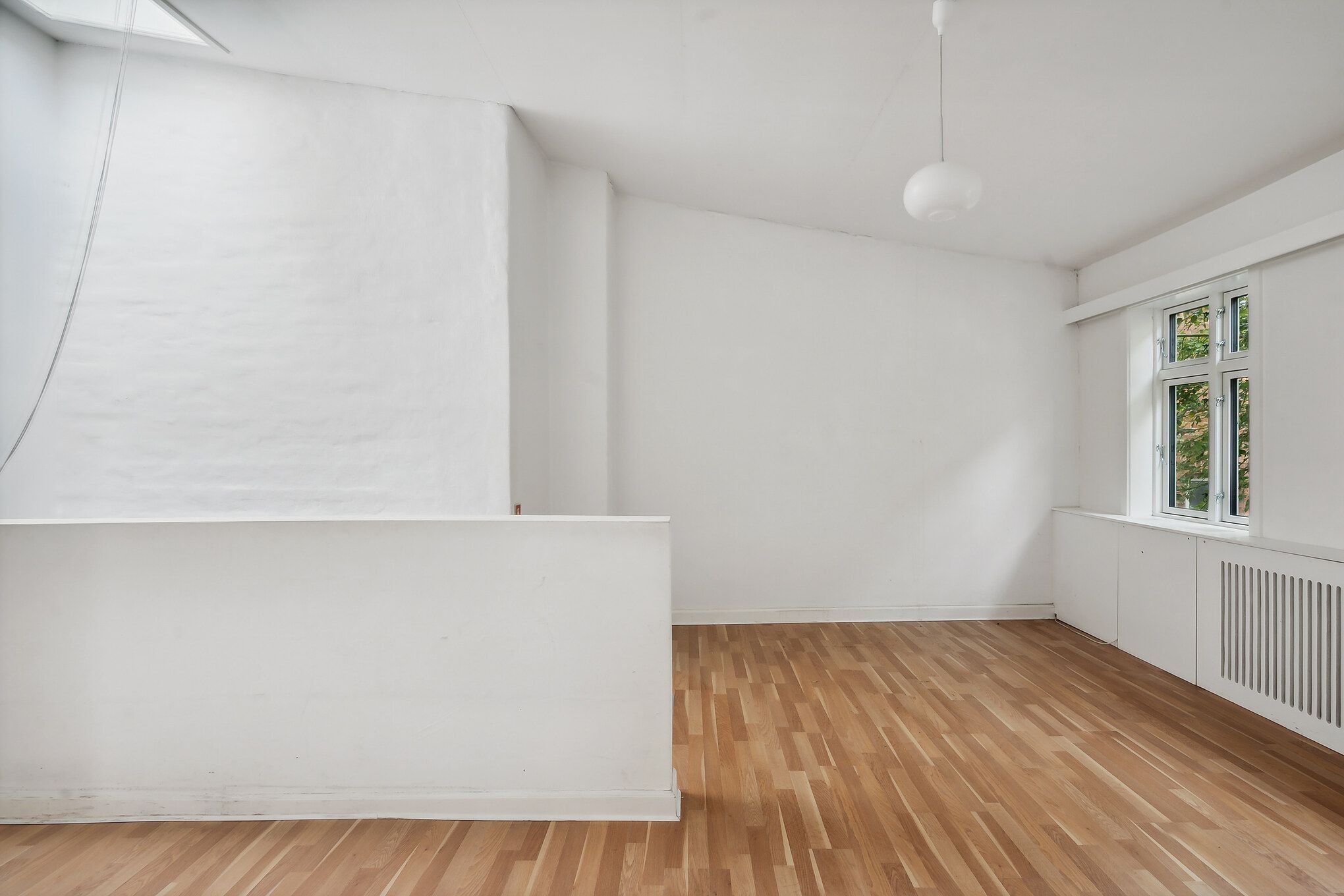 Lyst og højloftet lokale med hvide vægge og lyse trægulve samt store vinduer og en hvid radiator i hjørnet