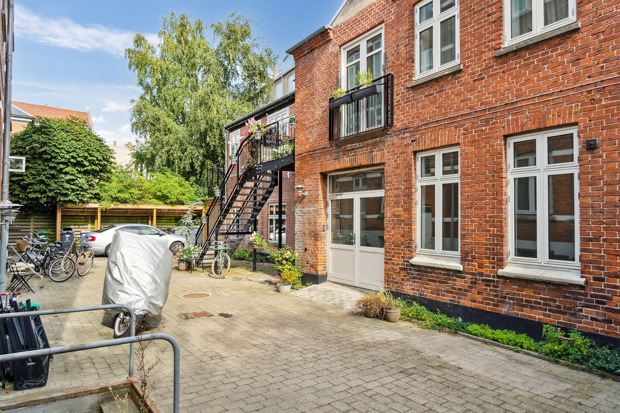 Rød murstensbygning med vinduer og balkoner, samt skur med cykler og bil, samt plantepotte og gårdhave med træer og grønne