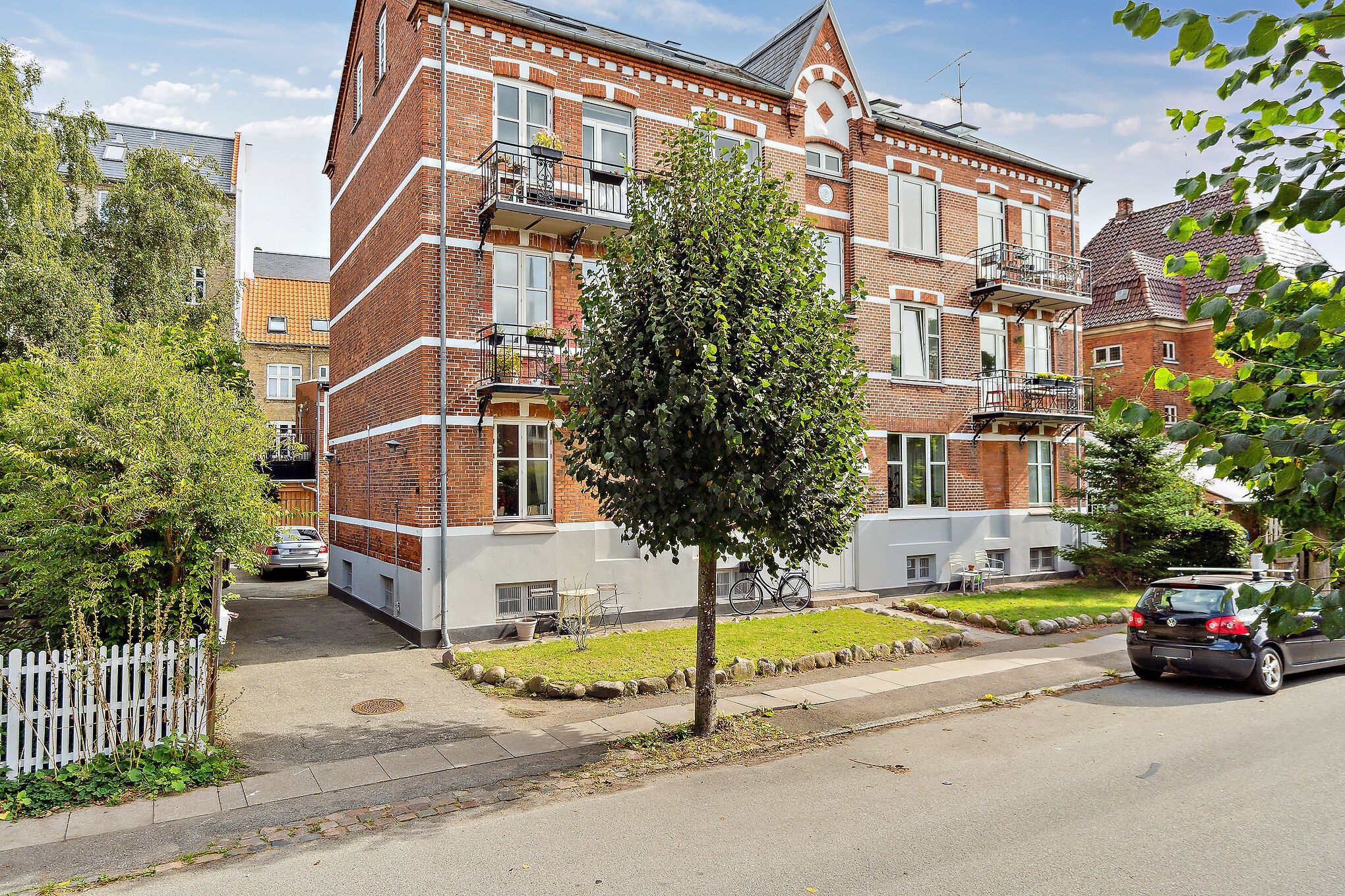 Et rødt murstenshus med hvide accenter og mange vinduer samt balkoner og haver. Et træ står foran huset, og en cykel er