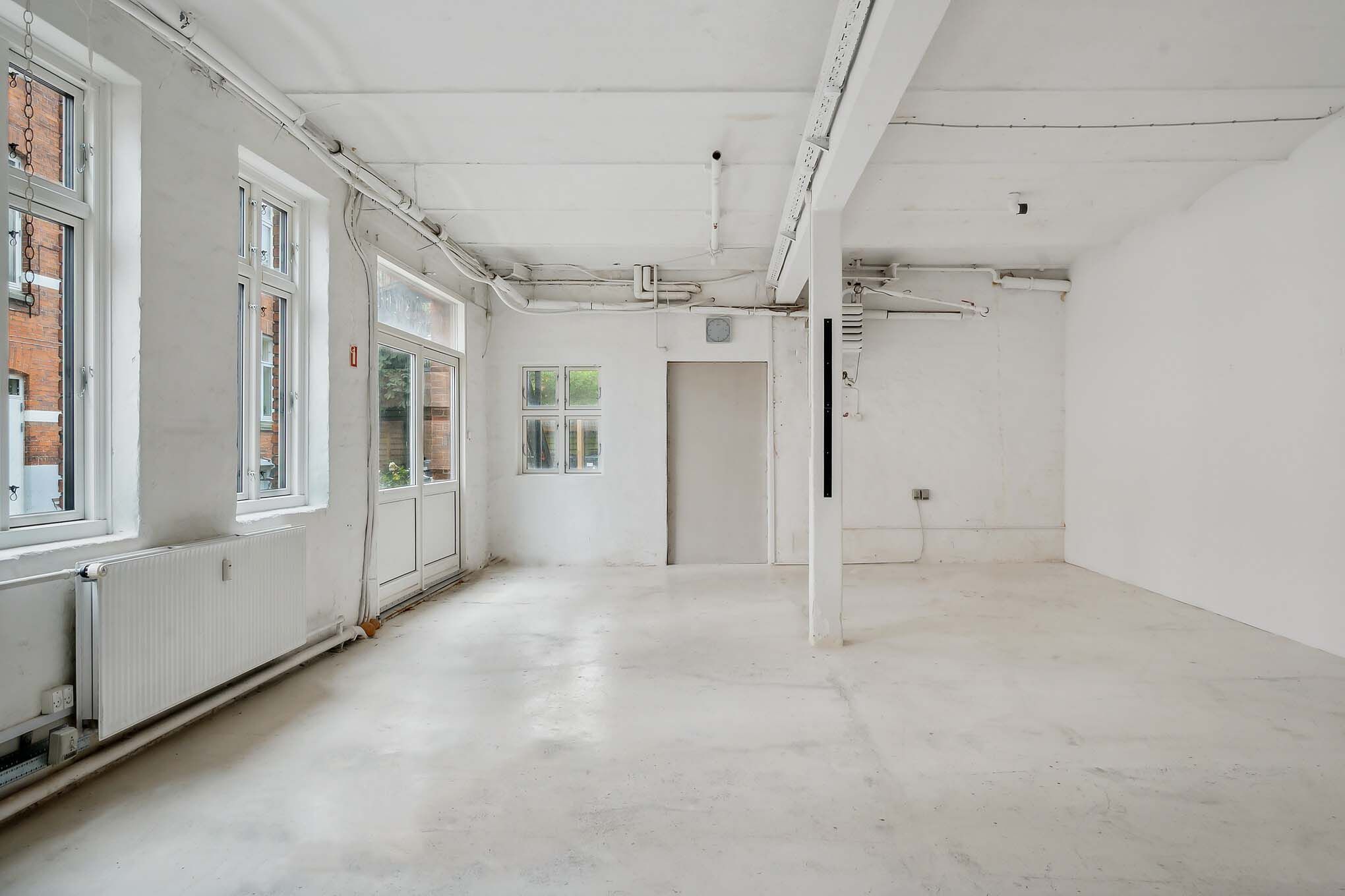 Kreativt værksted med højloft og hvide vægge, radiatorer, vinduer og døre, og lyse gulv.