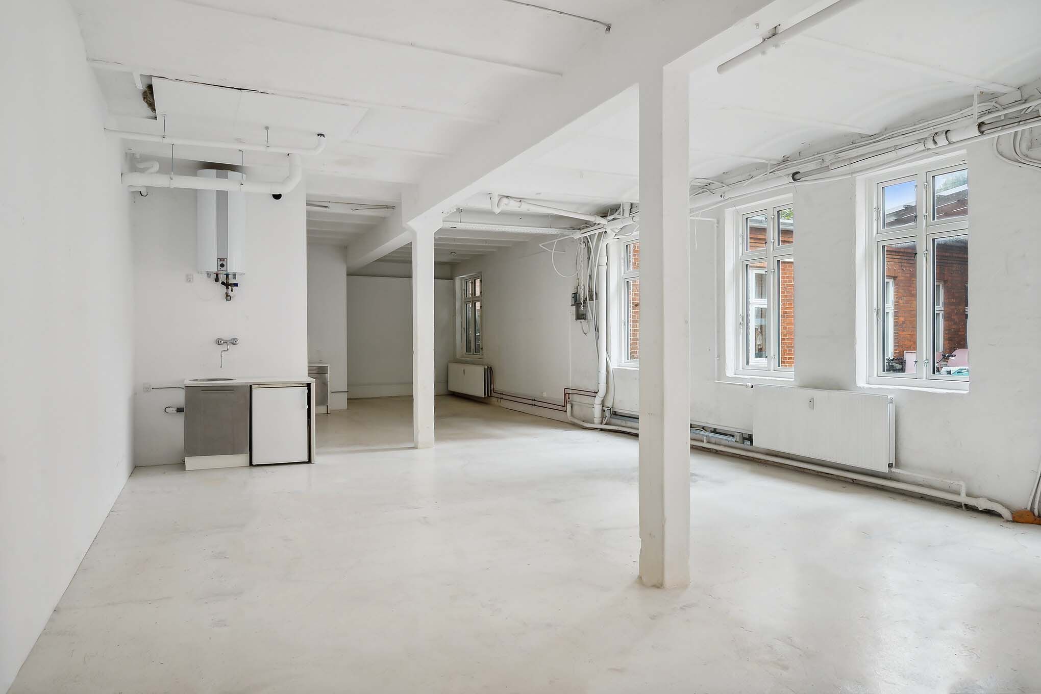 Lokale med højloft og store vinduer, indretning i hvidt, radiatorer og et lille køkkenhjørne med køleskab og vaskebækken