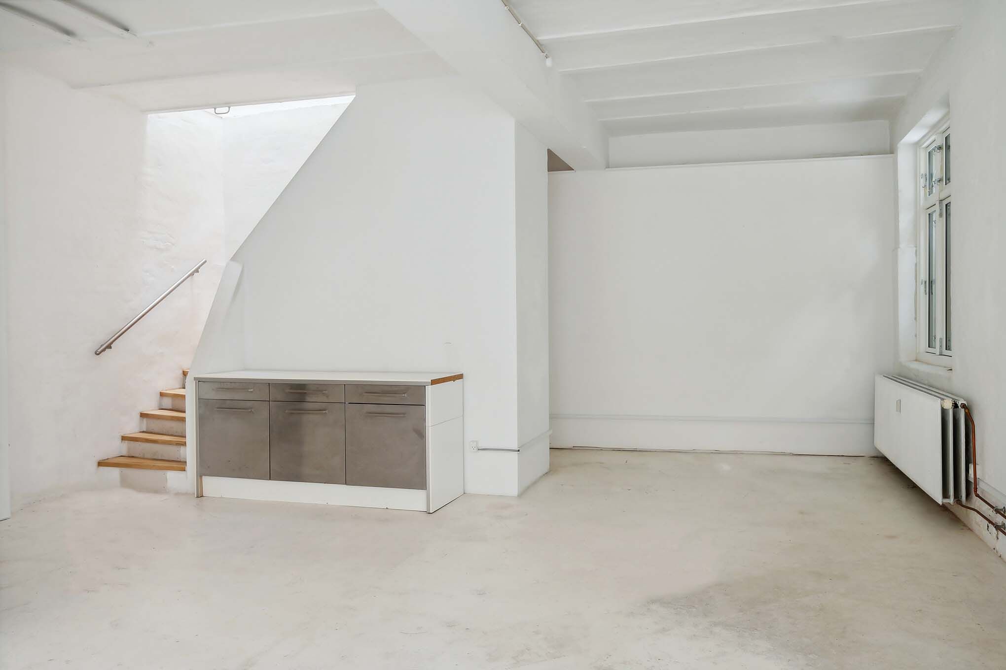 Lokale med hvide vægge og loft, lysegrå gulv, en trætrappe og et træbord med fire skuffer