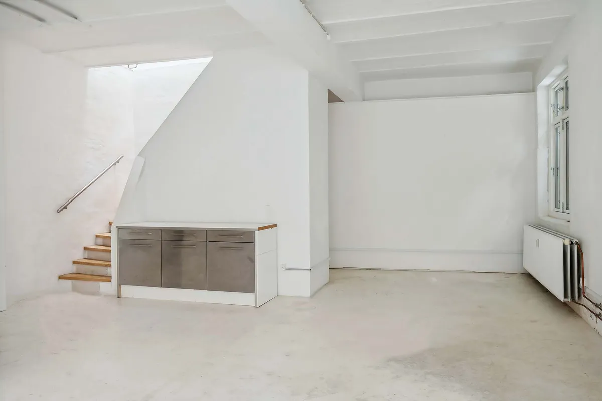 Lokale med hvide vægge og loft, lysegrå gulv, en trætrappe og et træbord med fire skuffer