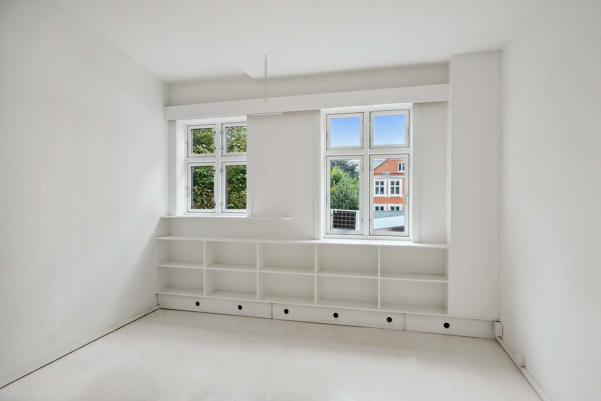 Minimalistisk, hvidt værelse med store vinduer og et indbygget højrekantet hyldesystem med flere indbygget rum