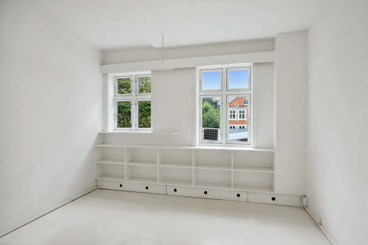 Minimalistisk, hvidt værelse med store vinduer og et indbygget højrekantet hyldesystem med flere indbygget rum