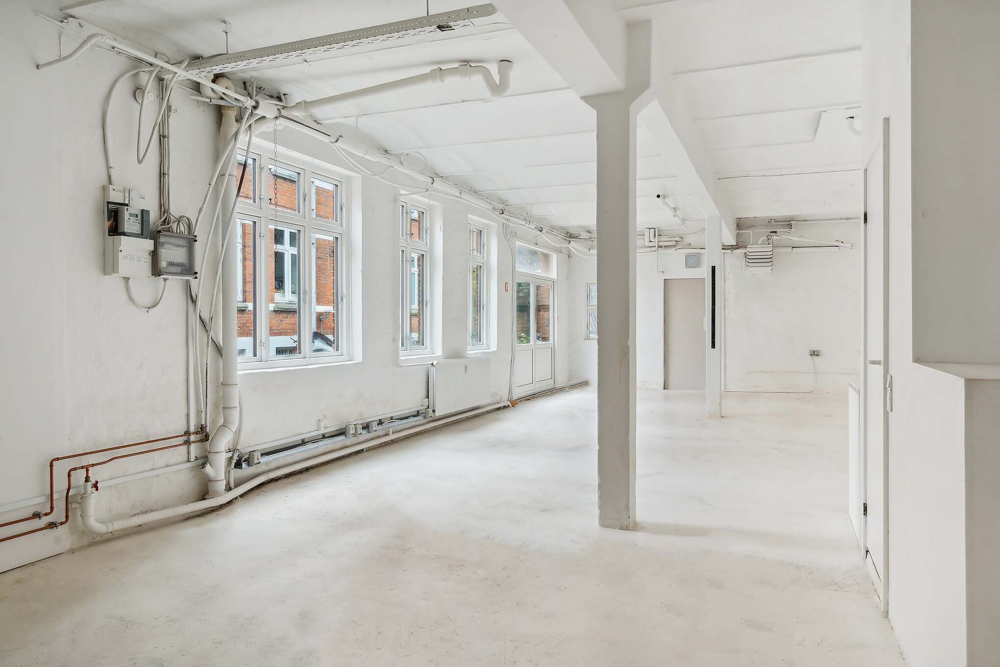 Lokale med hvide vægge og lofter, flere vinduer, rør og elektriske ledninger, hvide gulvbelægning og lysekroner
