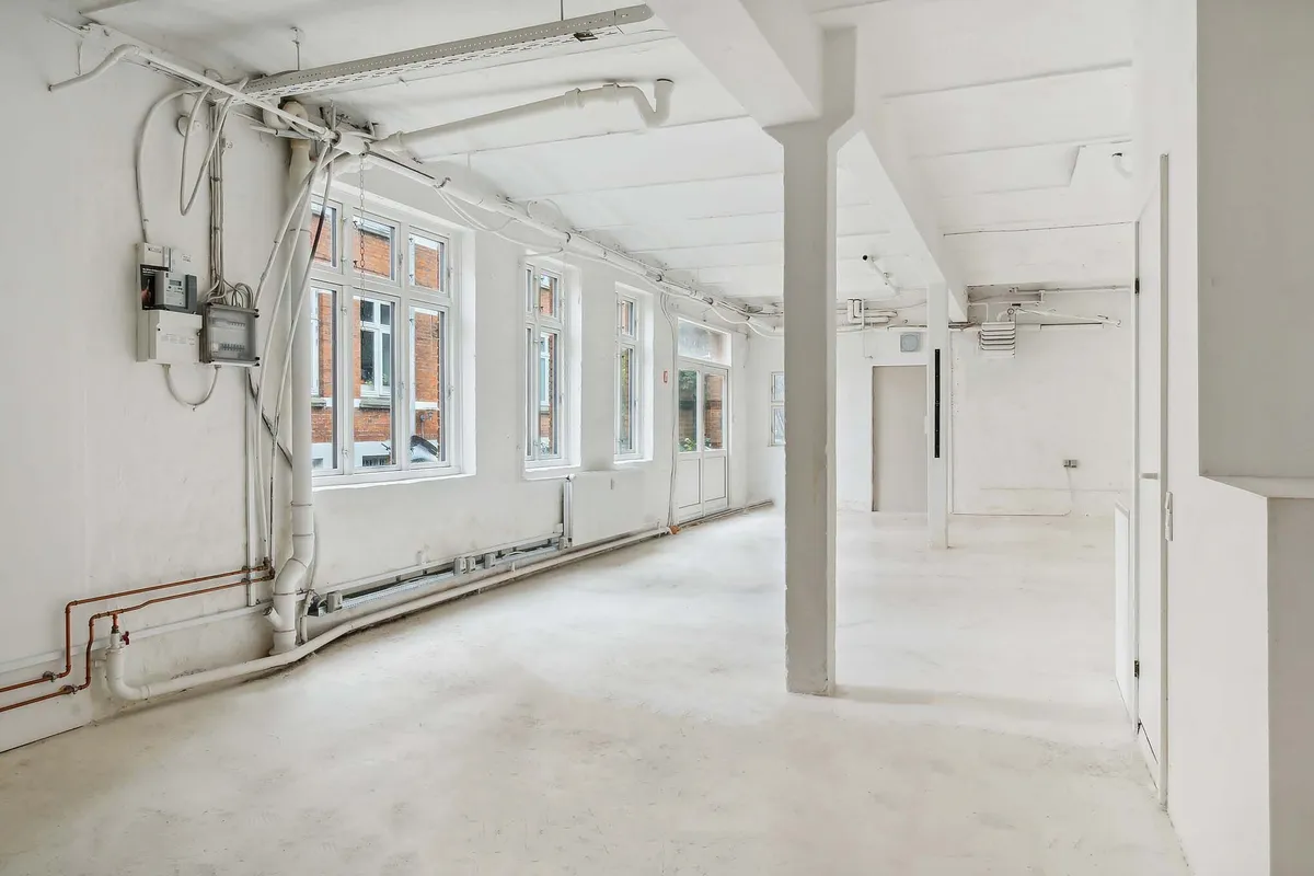 Lokale med hvide vægge og lofter, flere vinduer, rør og elektriske ledninger, hvide gulvbelægning og lysekroner
