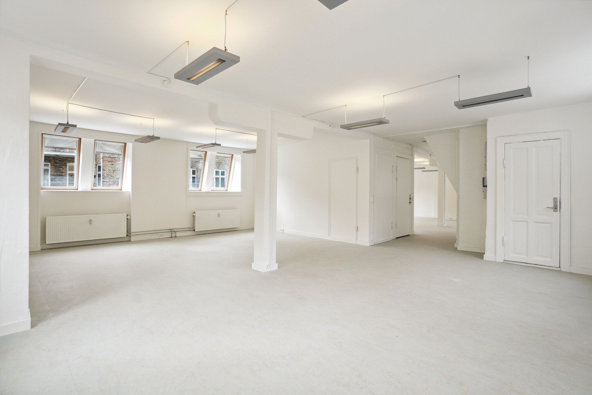 Lejlighed med store vinduer, hvidt loft og gulv, hvide vægge, radiatorer, to døre og indbyggede lysekroner