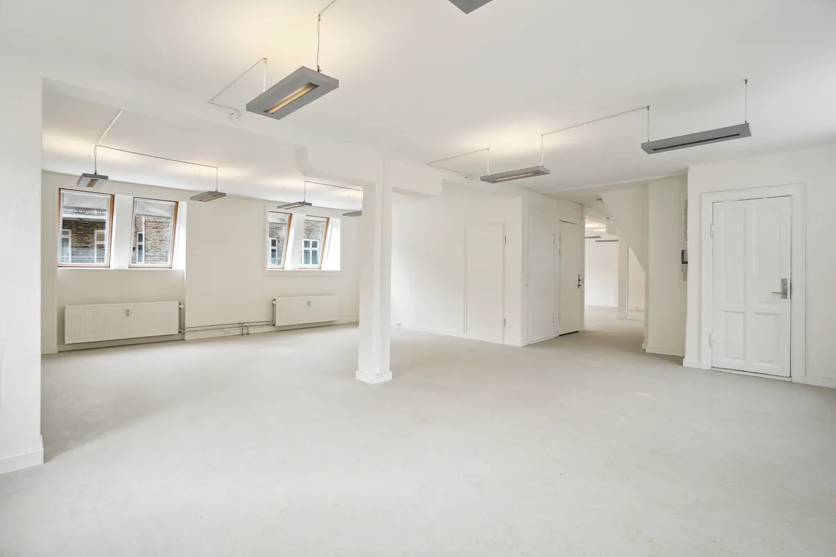 Lejlighed med store vinduer, hvidt loft og gulv, hvide vægge, radiatorer, to døre og indbyggede lysekroner