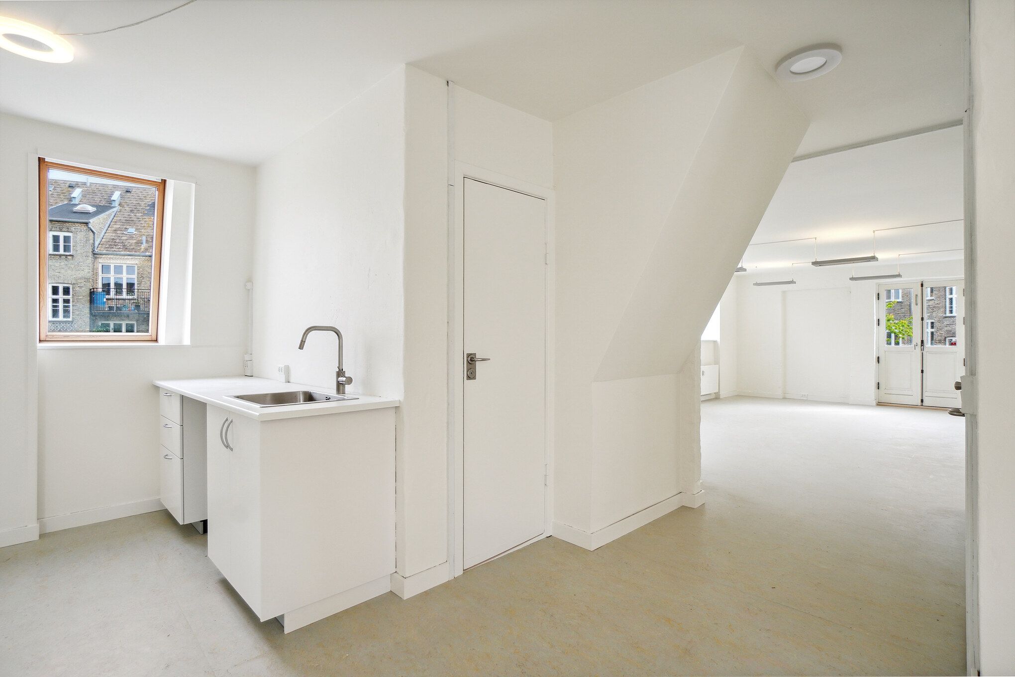 En tom stue med hvide vægge, loft og gulv og et køkken med lysegrå vask og hvidt skab