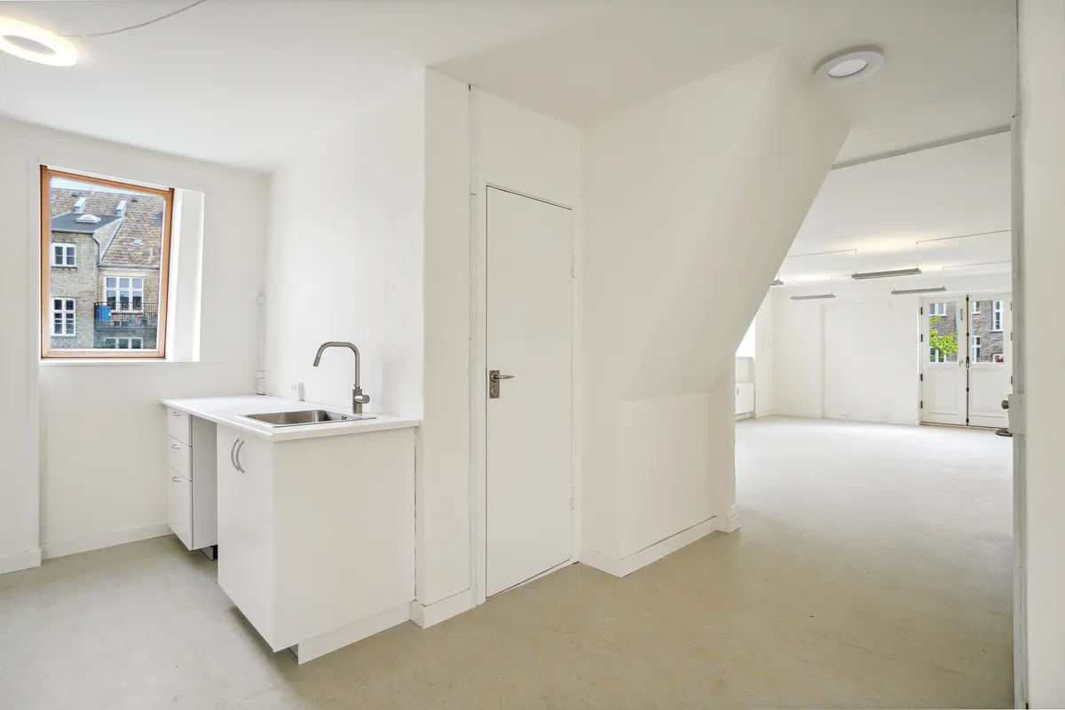 En tom stue med hvide vægge, loft og gulv og et køkken med lysegrå vask og hvidt skab